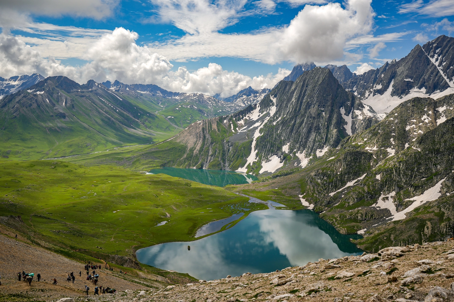 ERHOLUNG DE LUXE - Wandern mit Achtsamkeit Allgäu - Atemberaubender Blick auf zwei alpine Seen inmitten grüner Wiesen und schneebedeckter Himalaya-Gipfel, während Wanderer den rauen Himalaya-Pfad entlangwandern.